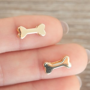 May include: Two small, gold-colored bone-shaped charms. The charms are likely made of metal and have a shiny finish. The bone charms are a popular design, often used in jewelry or craft projects.