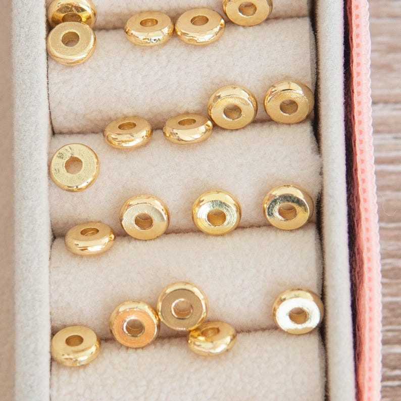 May include: A close-up of a pink jewelry box with multiple compartments filled with gold-colored round beads.