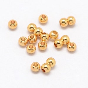May include: A scattering of small, shiny, gold-coloured beads on a white surface. Each bead is round with a hole through the centre, designed for stringing. The beads have a polished, reflective surface, suggesting a metallic material.