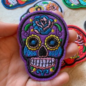 May include: Purple embroidered sugar skull patch with a blue and pink flower, yellow and blue eyes, and a black mouth.