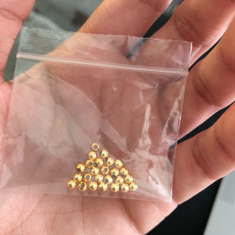 May include: A transparent plastic bag filled with small, shiny, gold-coloured beads. The beads are arranged in a triangular formation, and appear to be made of metal. These beads are likely used for jewellery making or other craft projects.