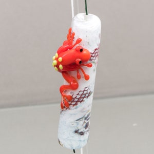 Puede incluir: Una figurita de rana de vidrio rojo posada sobre una cuenta de vidrio blanca y marrón. La cuenta tiene una superficie texturizada y un patrón de piel de serpiente.