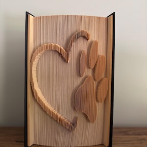 May include: A book sculpture featuring a heart and paw print design, created by folding the pages. The heart shape is on the left, and the paw print is on the right. The book has a black spine and is displayed on a wooden surface.