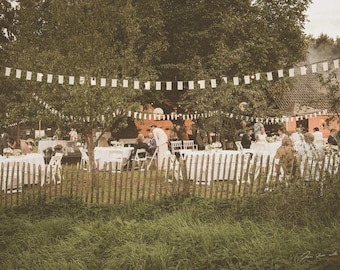 May include: An outdoor wedding reception with white tables and chairs set up under a canopy of trees. A string of white flags hangs above the tables, and guests are seated at the tables.