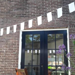May include: A brick building with a black door and window frames. White rectangular flags are strung across the top of the door. Purple flowers and green plants are in front of the building.
