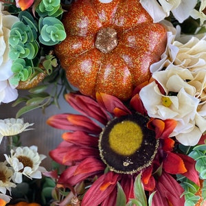 K&ouml;nnte beinhalten: Ein Herbstkranz mit einer roten Sonnenblume, wei&szlig;en Hortensien und kleinen K&uuml;rbissen mit goldenen Glitzer. Der Kranz ist mit gr&uuml;nen Bl&auml;ttern und wei&szlig;en Beeren verziert.