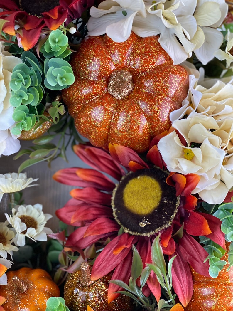 K&ouml;nnte beinhalten: Ein Herbstkranz mit einer roten Sonnenblume, wei&szlig;en Hortensien und kleinen K&uuml;rbissen mit goldenen Glitzer. Der Kranz ist mit gr&uuml;nen Bl&auml;ttern und wei&szlig;en Beeren verziert.