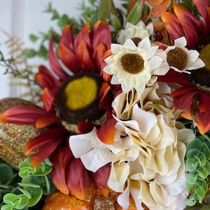 K&ouml;nnte beinhalten: Ein Herbstkranz mit einem K&uuml;rbis, Sonnenblumen und wei&szlig;en Blumen. Der Kranz ist mit Gr&uuml;n verziert und hat einen rustikalen Look.
