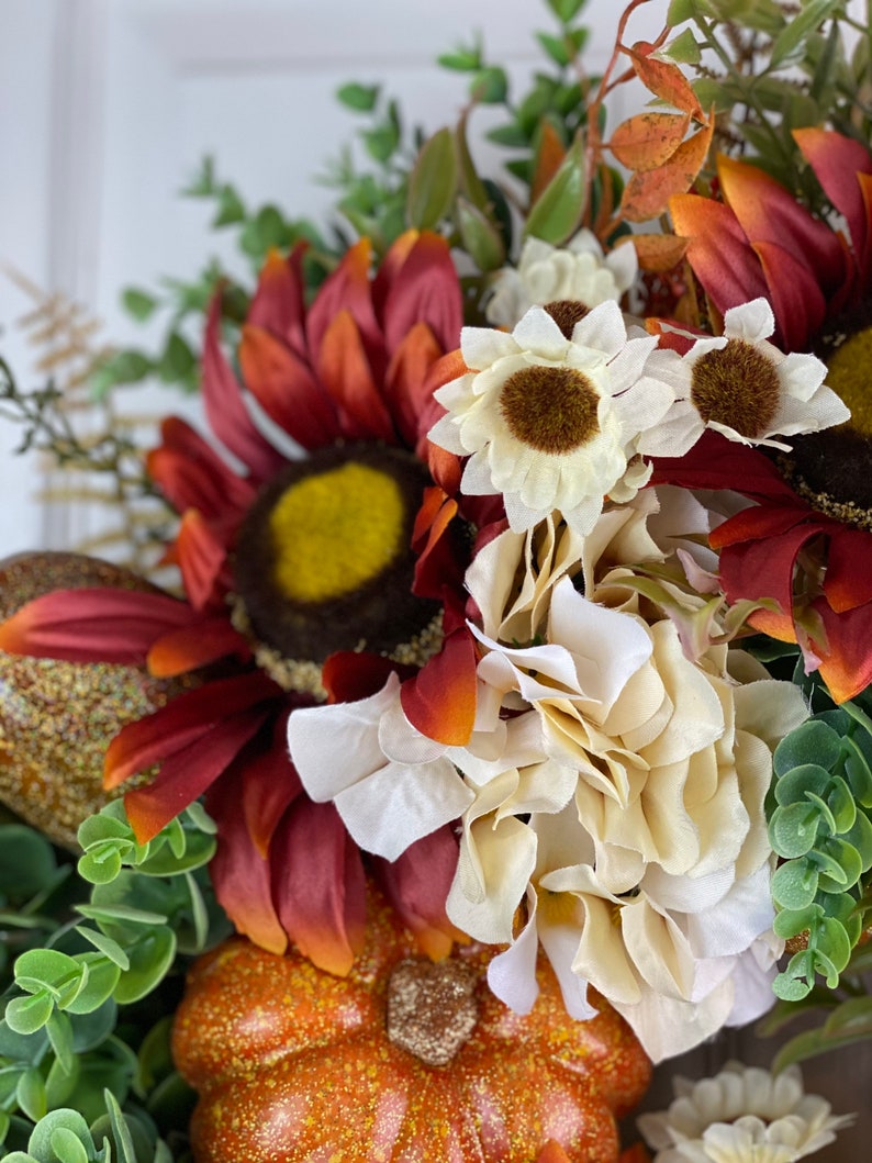 K&ouml;nnte beinhalten: Ein Herbstkranz mit einem K&uuml;rbis, Sonnenblumen und wei&szlig;en Blumen. Der Kranz ist mit Gr&uuml;n verziert und hat einen rustikalen Look.