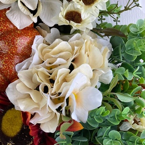 K&ouml;nnte beinhalten: Ein Herbstkranz mit verschiedenen k&uuml;nstlichen Blumen, darunter wei&szlig;e Hortensien, Sonnenblumen und Gr&uuml;n. Der Kranz ist mit zwei kleinen K&uuml;rbissen verziert.