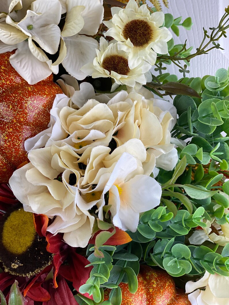 K&ouml;nnte beinhalten: Ein Herbstkranz mit verschiedenen k&uuml;nstlichen Blumen, darunter wei&szlig;e Hortensien, Sonnenblumen und Gr&uuml;n. Der Kranz ist mit zwei kleinen K&uuml;rbissen verziert.