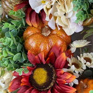 K&ouml;nnte beinhalten: Ein Herbstkranz mit einer roten Sonnenblume, wei&szlig;en Hortensien, gr&uuml;nen Bl&auml;ttern und zwei kleinen orangefarbenen K&uuml;rbissen mit Glitzerakzenten.