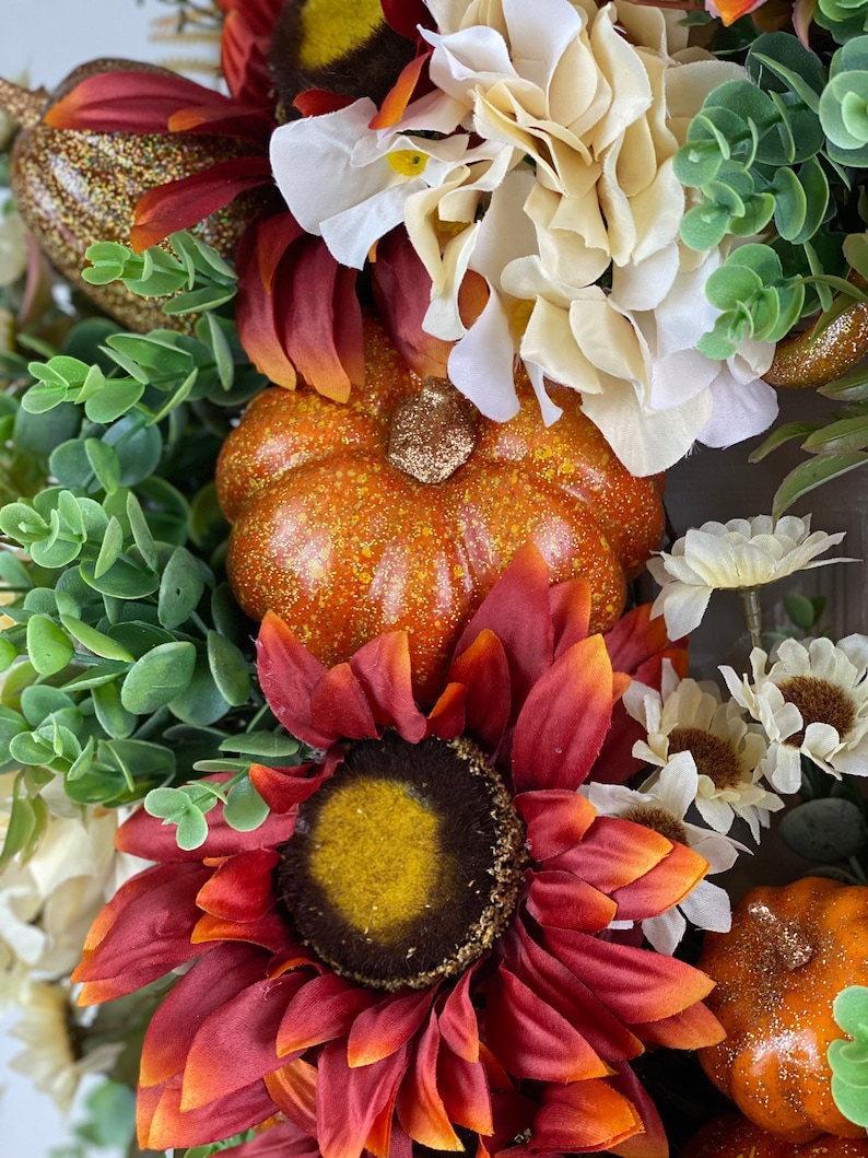 K&ouml;nnte beinhalten: Ein Herbstkranz mit einer roten Sonnenblume, wei&szlig;en Hortensien, gr&uuml;nen Bl&auml;ttern und zwei kleinen orangefarbenen K&uuml;rbissen mit Glitzerakzenten.