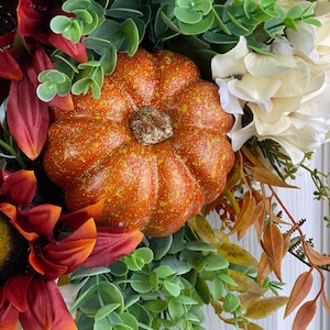 K&ouml;nnte beinhalten: Ein Herbstkranz mit einem kleinen orangefarbenen K&uuml;rbis, roten und wei&szlig;en Blumen und gr&uuml;nen Eukalyptusbl&auml;ttern.