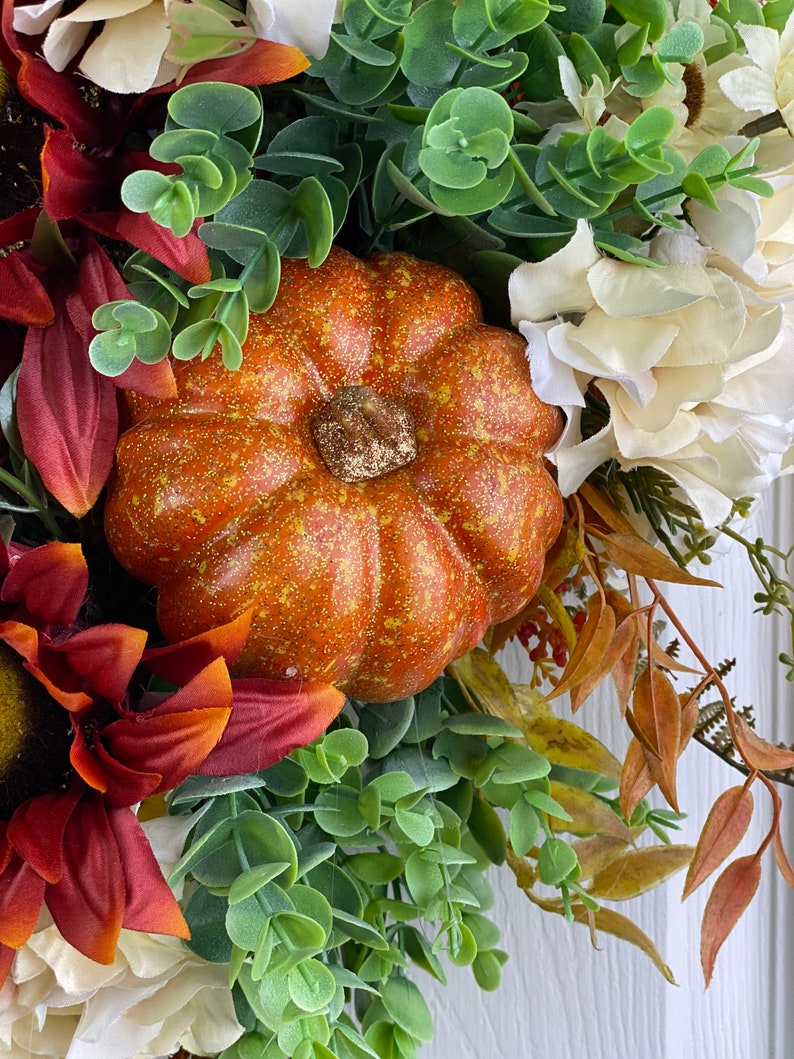 K&ouml;nnte beinhalten: Ein Herbstkranz mit einem kleinen orangefarbenen K&uuml;rbis, roten und wei&szlig;en Blumen und gr&uuml;nen Eukalyptusbl&auml;ttern.
