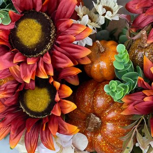 K&ouml;nnte beinhalten: Ein Herbstkranz mit roten und orangefarbenen Sonnenblumen, wei&szlig;en Bl&uuml;ten und kleinen orangefarbenen K&uuml;rbissen mit Glitzerakzenten.