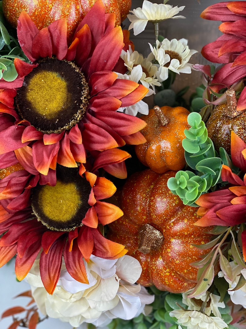 K&ouml;nnte beinhalten: Ein Herbstkranz mit roten und orangefarbenen Sonnenblumen, wei&szlig;en Bl&uuml;ten und kleinen orangefarbenen K&uuml;rbissen mit Glitzerakzenten.