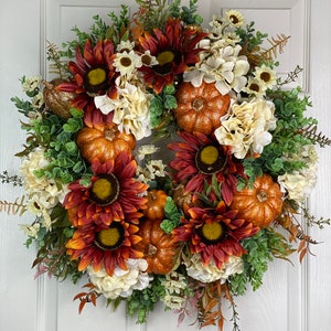 K&ouml;nnte beinhalten: Ein Herbstkranz mit orangefarbenen K&uuml;rbissen, Sonnenblumen und wei&szlig;en Hortensien. Der Kranz ist mit Gr&uuml;n verziert und hat einen rustikalen, l&auml;ndlichen Stil.
