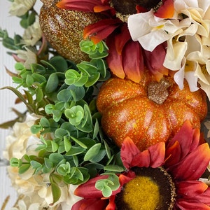 K&ouml;nnte beinhalten: Ein Herbstkranz mit roten und orangen Sonnenblumen, wei&szlig;en Hortensien, gr&uuml;nen Eukalyptusbl&auml;ttern und zwei kleinen K&uuml;rbissen.
