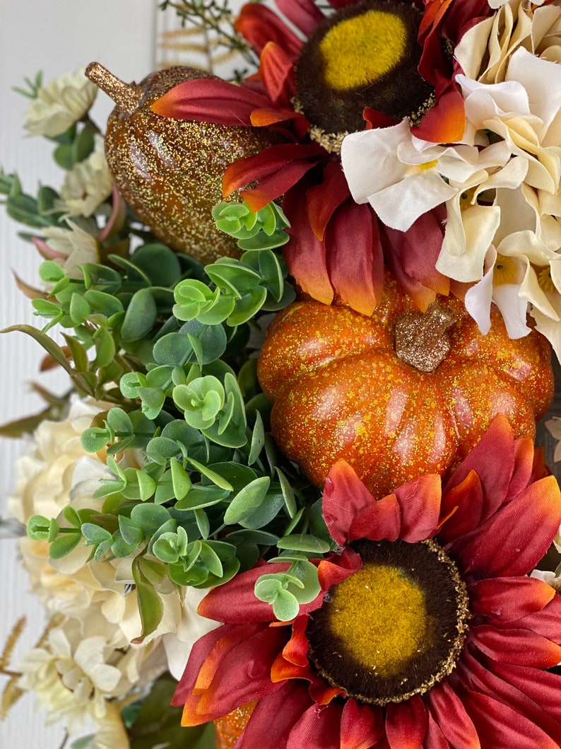 K&ouml;nnte beinhalten: Ein Herbstkranz mit roten und orangen Sonnenblumen, wei&szlig;en Hortensien, gr&uuml;nen Eukalyptusbl&auml;ttern und zwei kleinen K&uuml;rbissen.