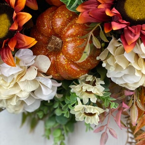 K&ouml;nnte beinhalten: Ein Herbstkranz mit orangefarbenen K&uuml;rbissen, roten und orangefarbenen Sonnenblumen und wei&szlig;en Hortensien. Der Kranz ist mit gr&uuml;nen Bl&auml;ttern und rosa Stielen verziert.