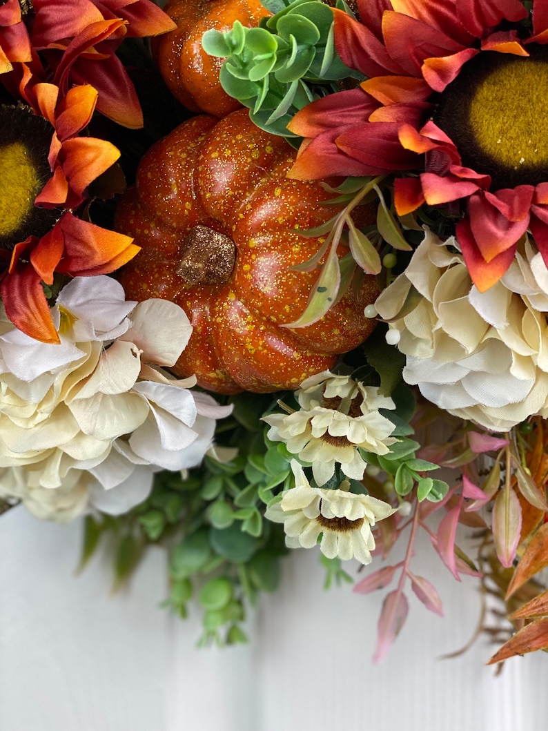 K&ouml;nnte beinhalten: Ein Herbstkranz mit orangefarbenen K&uuml;rbissen, roten und orangefarbenen Sonnenblumen und wei&szlig;en Hortensien. Der Kranz ist mit gr&uuml;nen Bl&auml;ttern und rosa Stielen verziert.