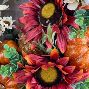 K&ouml;nnte beinhalten: Ein Herbstkranz mit zwei roten Sonnenblumen, orangefarbenen K&uuml;rbissen und wei&szlig;en Blumen. Die Sonnenblumen haben eine dunkelbraune Mitte mit einer gelben Mitte und die K&uuml;rbisse haben eine glitzernde orangefarbene Oberfl&auml;che.