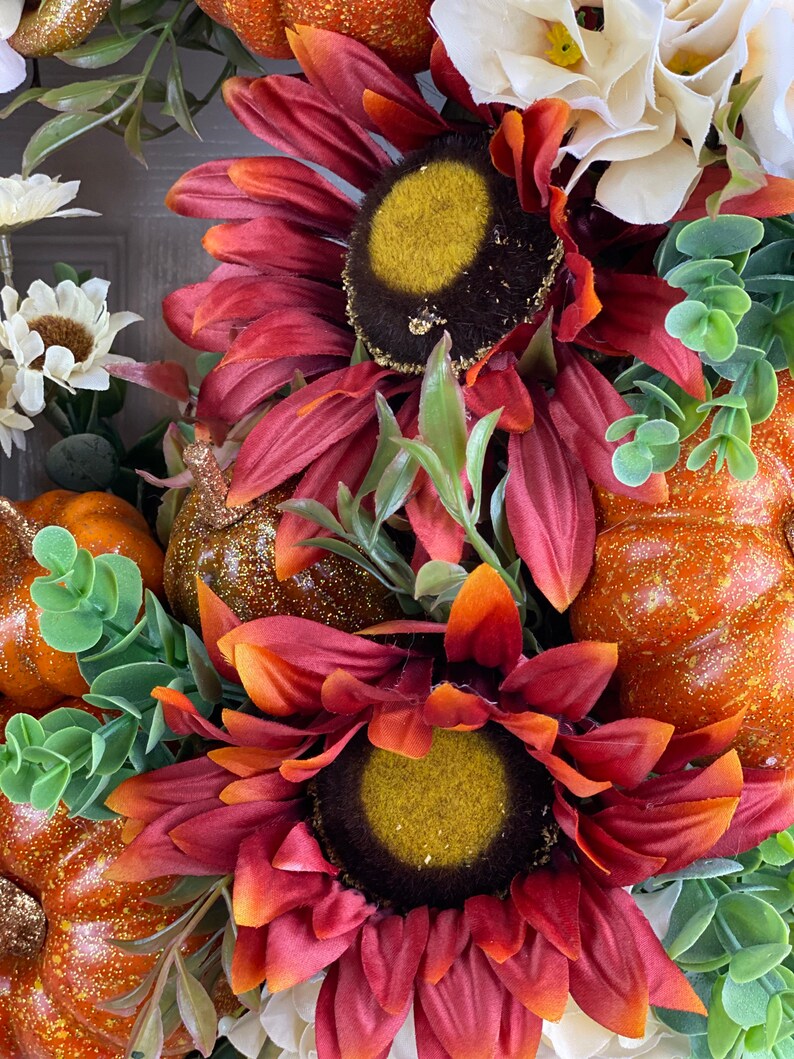 K&ouml;nnte beinhalten: Ein Herbstkranz mit zwei roten Sonnenblumen, orangefarbenen K&uuml;rbissen und wei&szlig;en Blumen. Die Sonnenblumen haben eine dunkelbraune Mitte mit einer gelben Mitte und die K&uuml;rbisse haben eine glitzernde orangefarbene Oberfl&auml;che.