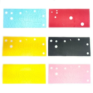 Puede incluir: Seis hojas de papel de colores con plantillas de alfabeto recortadas. Las hojas son azules, rojas, amarillas, negras, rosas y amarillas. Cada hoja tiene un color diferente y una plantilla de alfabeto diferente.