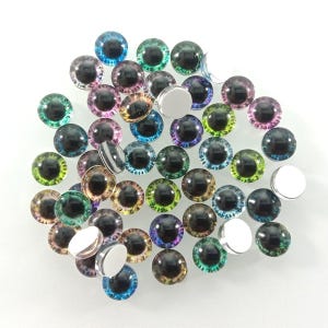 May include: A selection of colourful glass doll eyes in shades of blue, green, purple, and yellow. Each eye features a black pupil and a reflective, domed surface. Several silver-coloured metal backings are also present.
