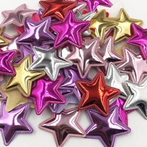 May include: A pile of colorful, metallic star-shaped embellishments. The stars are made of a shiny, textured material and come in a variety of colors, including gold, silver, pink, purple, and red.