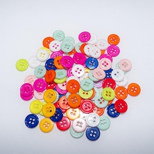 Puede incluir: Un gran surtido de botones de plástico coloridos con cuatro agujeros cada uno. Los botones son de varios tonos de rosa, azul, verde, amarillo, naranja y blanco.
