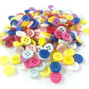 Puede incluir: Un montón grande de botones de plástico de colores en varios tonos de rojo, azul, amarillo, rosa y blanco. Los botones tienen cuatro agujeros y están dispuestos en un patrón aleatorio.