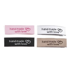 Puede incluir: Cuatro etiquetas de tela con el texto "hand made with love" y un diseño de corazón. Las etiquetas son de color rosa, blanco, negro y marrón.