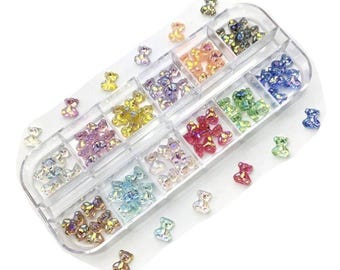 May include: A clear plastic container filled with small iridescent bear-shaped charms in various colors, including pink, blue, green, yellow, and red. The charms are arranged in rows in the container, and there are some loose charms scattered around the container.