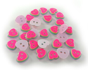 May include: A collection of pink heart-shaped buttons with rainbow layered designs. The buttons are made of plastic and have two holes for sewing.