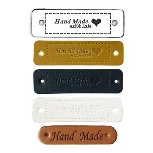Puede incluir: Cinco etiquetas de cuero con el texto "Hand Made" y un corazón. Las etiquetas son de diferentes colores: blanco, negro, marrón, beige y marrón claro.