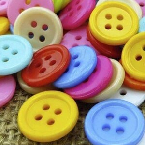 Puede incluir: Una colección de botones coloridos en varios tonos de rosa, amarillo, rojo, azul y crema. Los botones son redondos con cuatro agujeros cada uno, y están dispersos sobre una superficie texturizada.