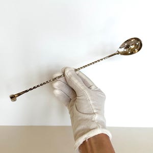 May include: A silver bar spoon with a twisted handle and a teardrop-shaped bowl. The spoon is held by a gloved hand against a white background. The spoon's length is approximately 12 inches.