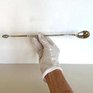May include: A silver bar spoon with a twisted handle and a teardrop-shaped bowl. The spoon has a decorative end with a small, ornate design. The spoon is held by a gloved hand against a white background.