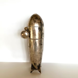 May include: A vintage silver cocktail shaker with a unique bullet or rocket shape. The shaker has a rounded top, a mid-section with a small handle, and a base with three fins. The metal has a tarnished, aged appearance.