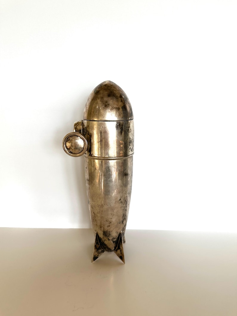 May include: A vintage silver cocktail shaker with a unique bullet or rocket shape. The shaker has a rounded top, a mid-section with a small handle, and a base with three fins. The metal has a tarnished, aged appearance.