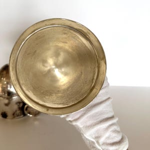 May include: A tarnished brass-colored bowl is held by a gloved hand. The bowl has a wide, shallow shape with a rounded bottom and a slightly raised rim. Another similar bowl is partially visible in the background.