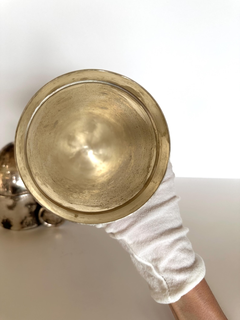 May include: A tarnished brass-colored bowl is held by a gloved hand. The bowl has a wide, shallow shape with a rounded bottom and a slightly raised rim. Another similar bowl is partially visible in the background.