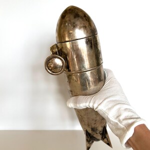 May include: A vintage silver-toned cocktail shaker with a bullet-like design. The shaker has a rounded top, a cylindrical body, and a small, circular handle. The bottom features two triangular fins. The shaker is held by a gloved hand.