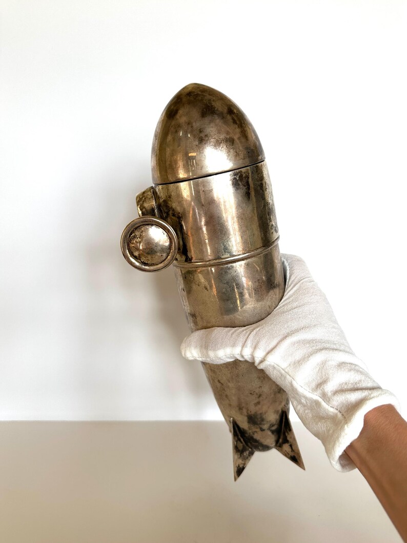 May include: A vintage silver-toned cocktail shaker with a bullet-like design. The shaker has a rounded top, a cylindrical body, and a small, circular handle. The bottom features two triangular fins. The shaker is held by a gloved hand.