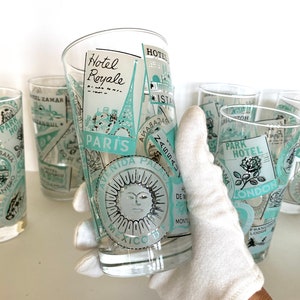 May include: Set of six clear glass tumblers with a turquoise and black design featuring travel-themed illustrations and text, including "Hotel Royale", "Paris", "Park Hotel", "London", "Avenida Palace", and "Mexico D.F."
