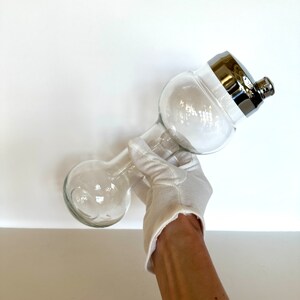 May include: A clear glass cocktail shaker with a bulbous base and a rounded top section. The shaker features a polished silver-colored metal cap with a small, threaded spout. The glass body is transparent, showing a smooth, curved silhouette.