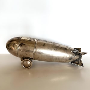 May include: A silver-colored, vintage-style cocktail shaker shaped like a zeppelin. The shaker has a cylindrical body with a rounded front and a finned tail. It features a small wheel and is likely made of metal.
