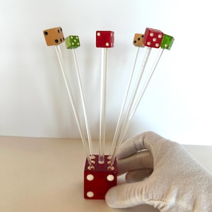 May include: A set of six dice-themed cocktail picks. Each pick features a small die in various colors: red, green, and tan, with white dot markings. The dice are mounted on clear, slender stems, and are held in a red die-shaped holder.